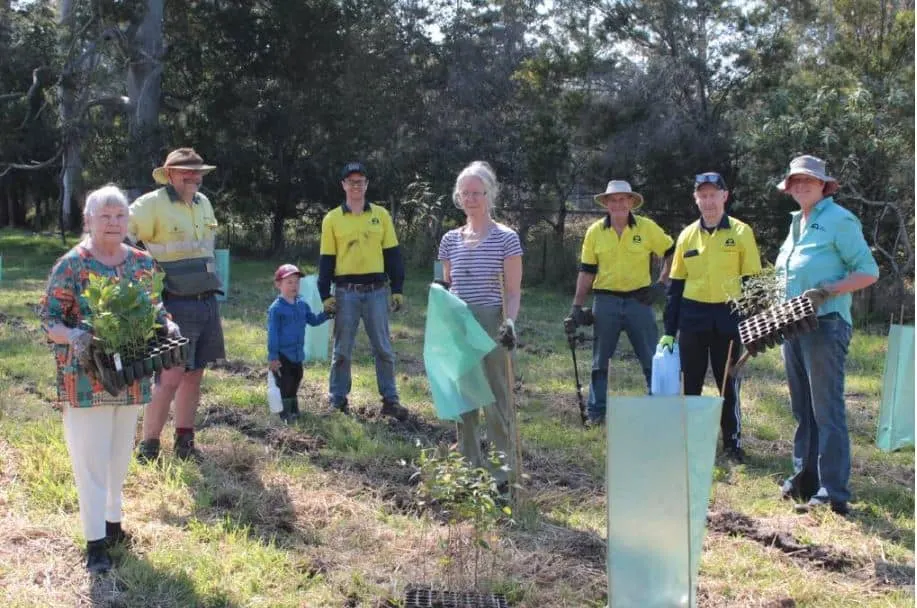 Volunteers and landholder in hi-vis and hats holding seedlings and tools beside tree guards in a paddock