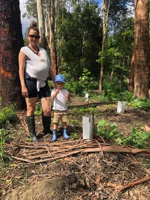Woman carrying a baby and a toddler in a cap standing by a tree guard on a bushland planting site