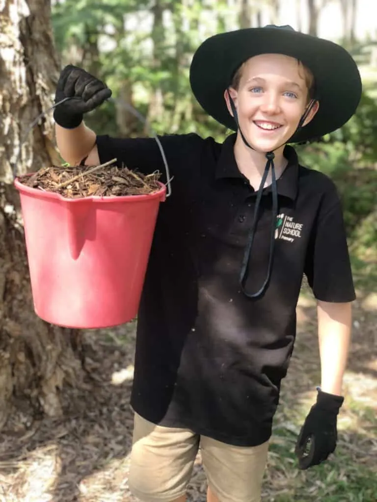 Child in a bush hat and gloves holding a red bucket of mulch beside a tree at a Nature School planting