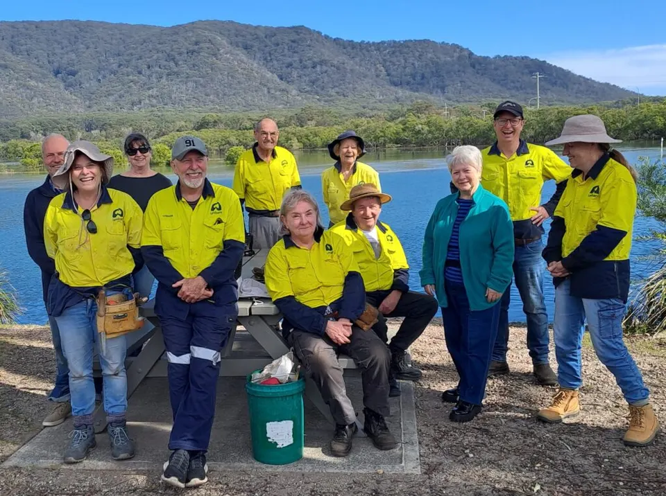 Landcare volunteers in yellow hi-vis shirts seated and standing together by a river with mountains behind